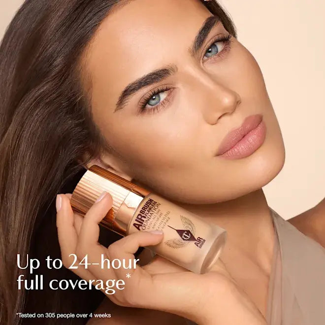 A close-up beauty ad showing a woman holding a bottle of Charlotte Tilbury airbrush foundation. The image features a warm beige background, promotional text reading 'Up to 24-hour full coverage*', and the foundation bottle has a metallic rose gold cap and frosted beige body.
