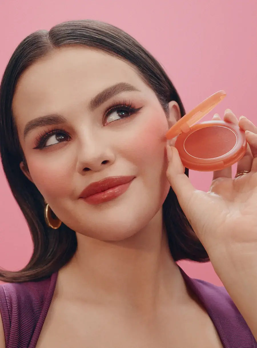 Rare Beauty Soft Pinch Cheek & Lip Trio Set Woman applying blush to her cheek with a pink background | MANSH