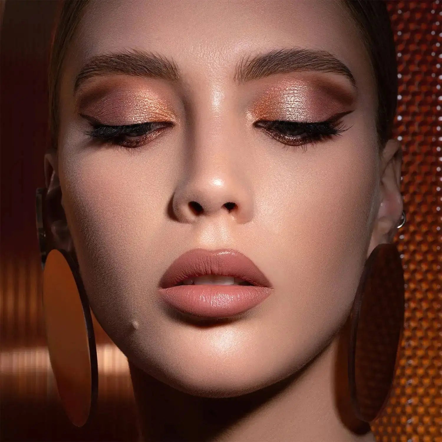 Close-up of a woman's face with NATASHA DENONA I Need a Warm Eyeshadow Palette makeup, wearing large earrings against a textured brown background