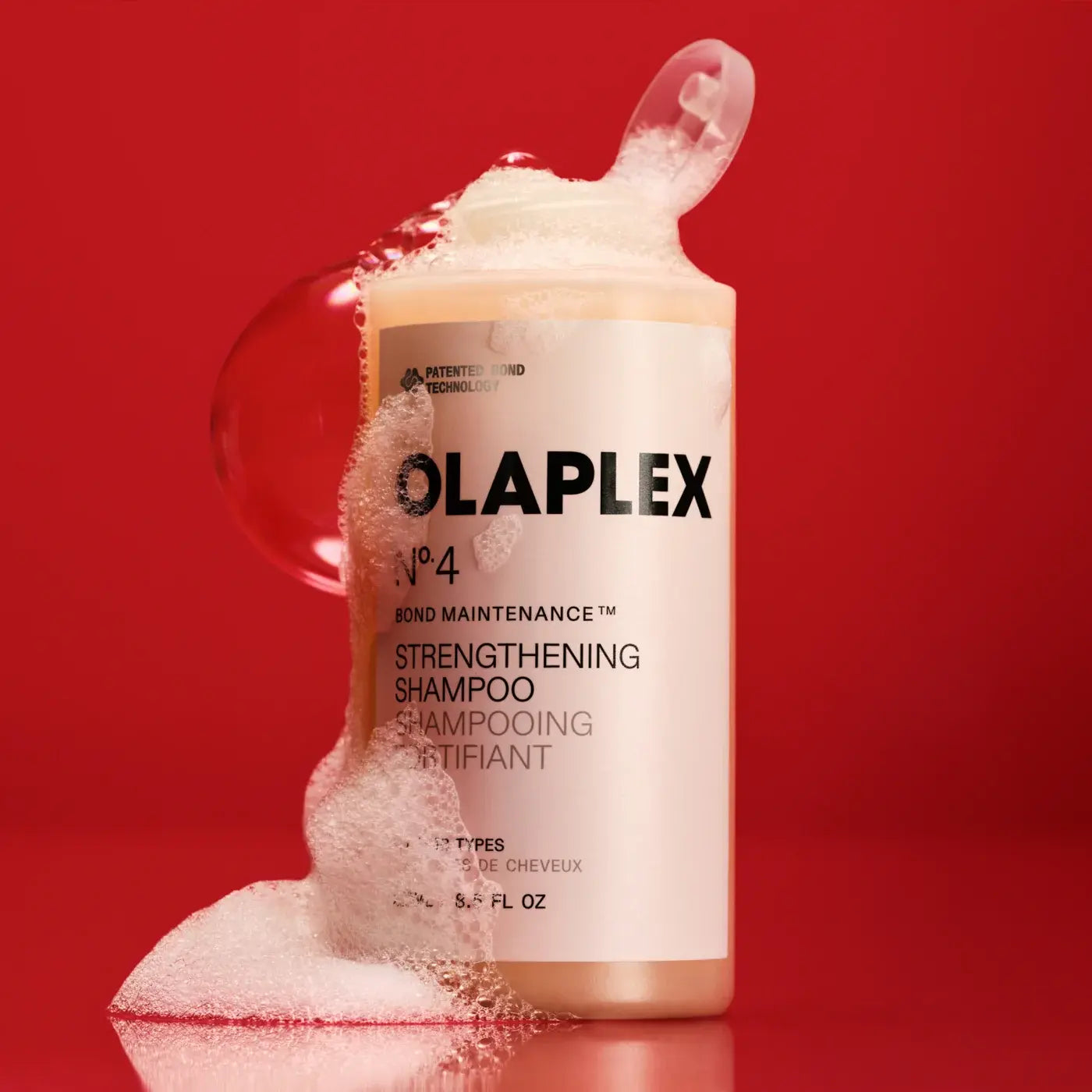 A bottle of Olaplex No. 4 Bond Maintenance Strengthening Shampoo with thick white foam overflowing from the top and pooling around the base, set against a solid red background.